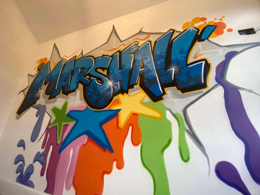 A bedroom wall spraypainted with the name Marshall in a graffiti style in blues with stars and splashes of bright colours below